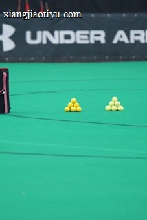 A sports field features a black and pink sports bag, two pyramids of yellow balls, and a portion of a person in dark athletic gear. The background displays a banner with the Under Armour logo.