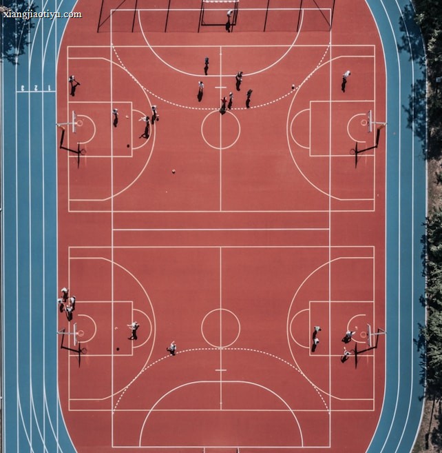 An aerial view of a large, vibrant sports complex featuring a red basketball court with multiple hoops and a surrounding blue running track. Several people are scattered across the area, engaged in various activities.