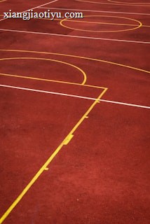 A sports court with a reddish-brown surface and bright yellow and white lines. The lines form geometric shapes, including rectangles and circles, suggesting markings for games like basketball or netball.